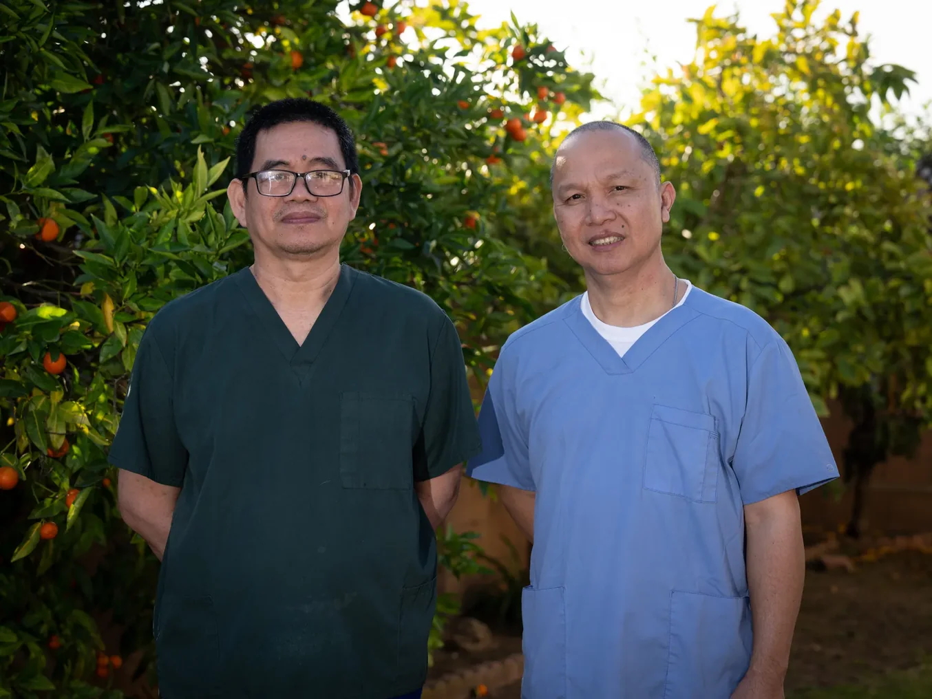 Two smiling male caregivers in medical scrubs standing in an outdoor orchard setting